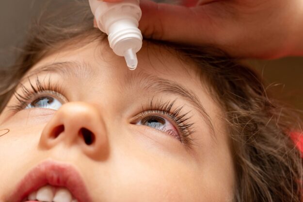 car de um menino com os olhos em destaque, um dos olhos vermelho, com um medicamento de gotas a ser colocado no olho inflamado